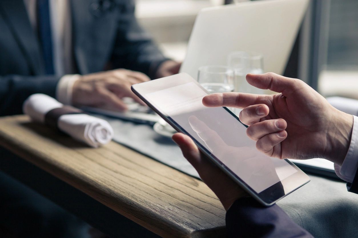 Close up of arms of two mature businessmen meeting in restaurant. They are sitting at the table. Man is using a laptop. His colleague is holding a tablet and pointing finger at it. Focus on his hands (Close up of arms of two mature businessmen meeting